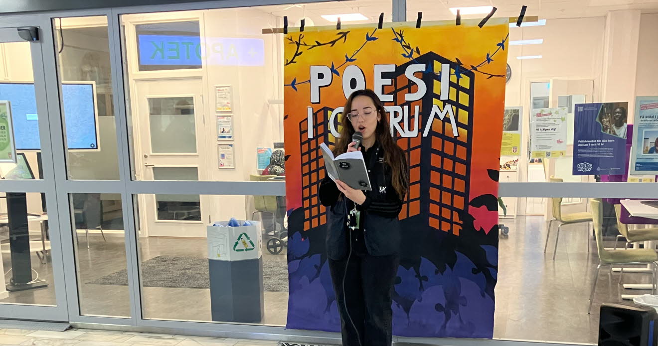 En ung kvinna står med en mikrofon i ett köpcentrum och läser ur en bok.