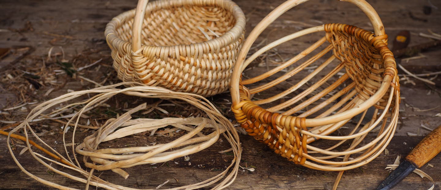 Två flätade korgar i naturmaterial, omgivna av rötter och en kniv på ett träbord.