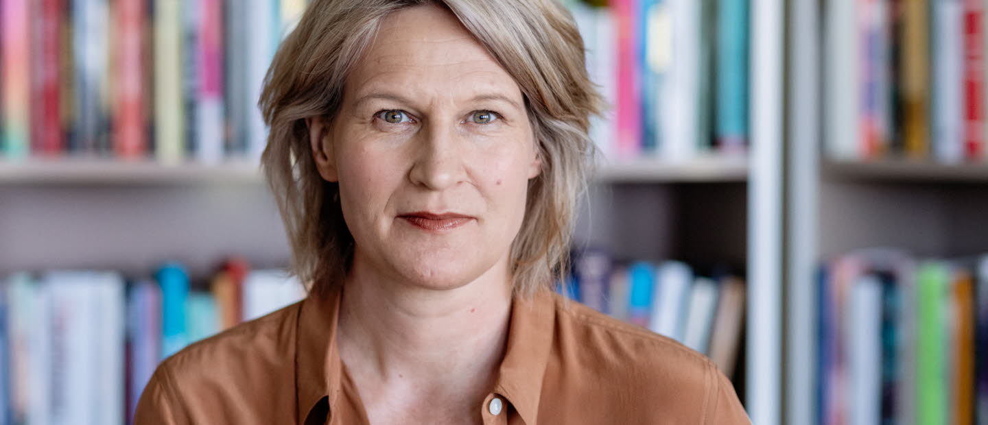 Medelålderskvinna i halvfigur. I bakgrunden bokhyllor fyllda med böcker. 