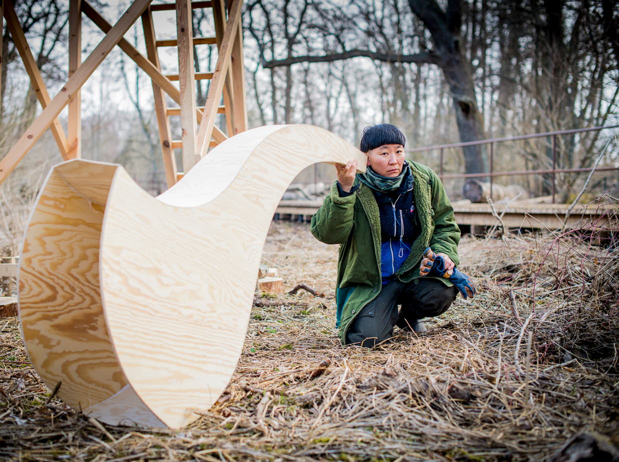 En kvinna som sitter utomhus lyssnar i en överdimensionerad tratt gjord av trä.