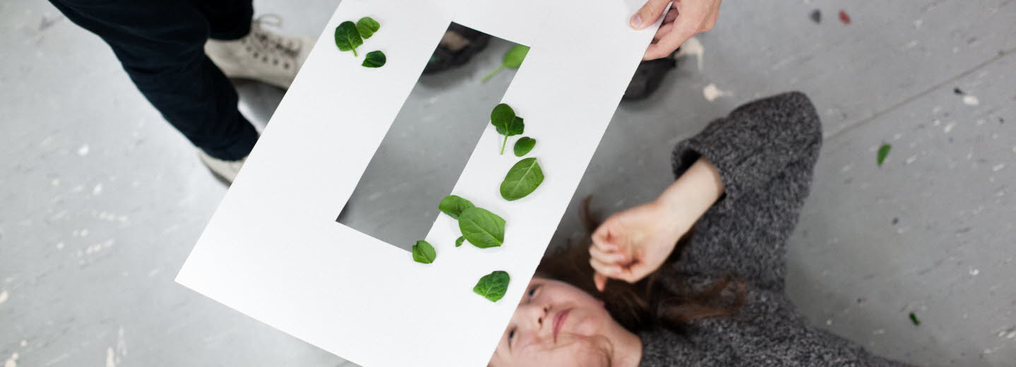 En kvinna ligger på rygg och tittar upp mot ett genombrutet papperark som hålls ovanför hennes huvud. Det ligger gröna blad på pappersarket. 