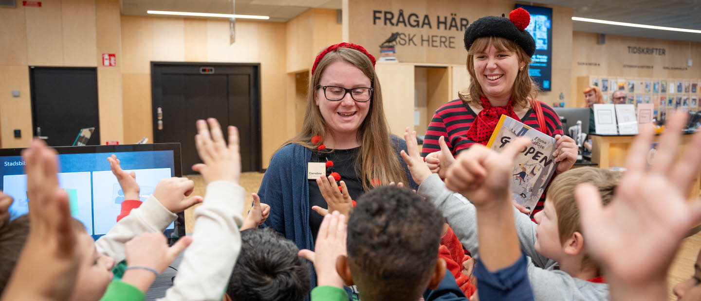 Två kvinnor står vända mot kameran i biblioteksmiljö. Framför dem, med ryggen mot kameran räcker många små barn upp händerna. 