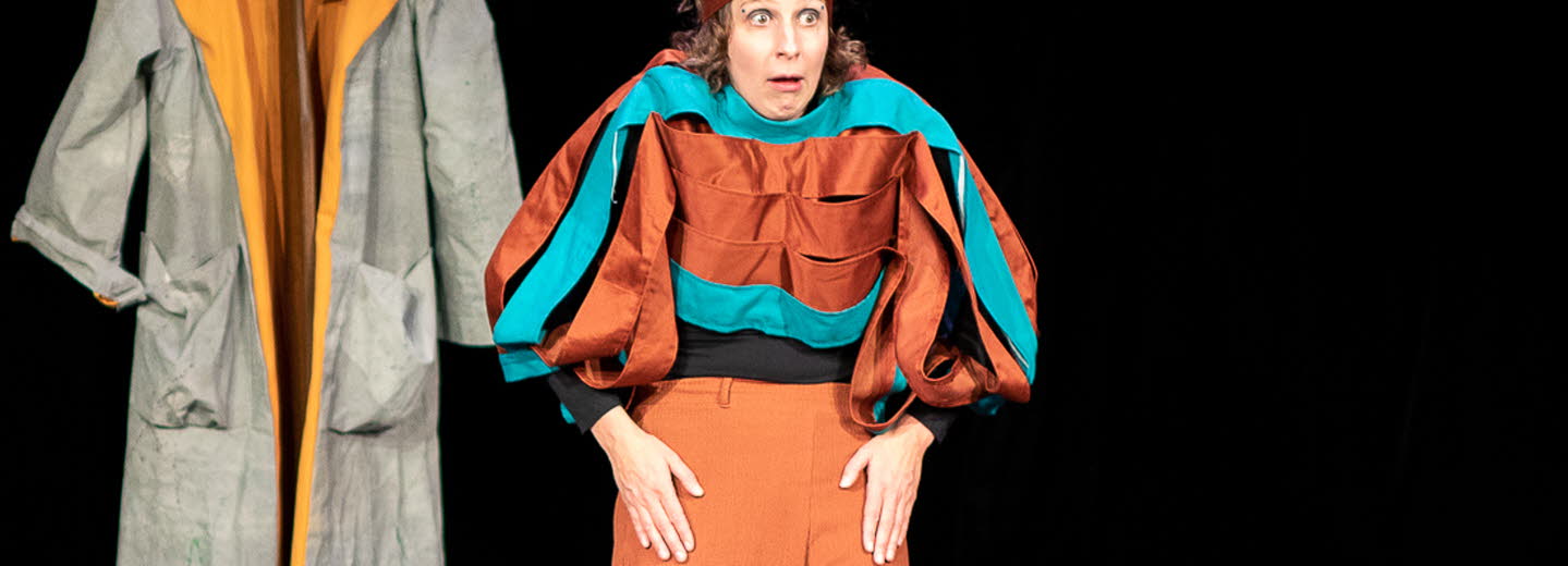 Två kvinnor på scen. En spelar på keyboard som ligger på ett figurliknande träbord och den andra tittar storögt mot publiken, axlarna är upphöjda. Båda har färgstarka kläder i turkost och ockra. I bakgrunden hänger en grå rock med tillhörande hatt.