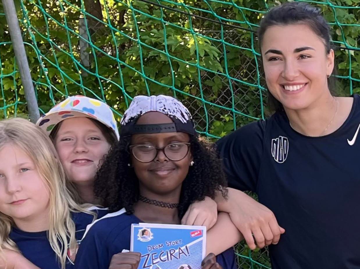 Barn i fotbollskläder som poserar tillsammans med en kvinna i ett fotbollsmål.