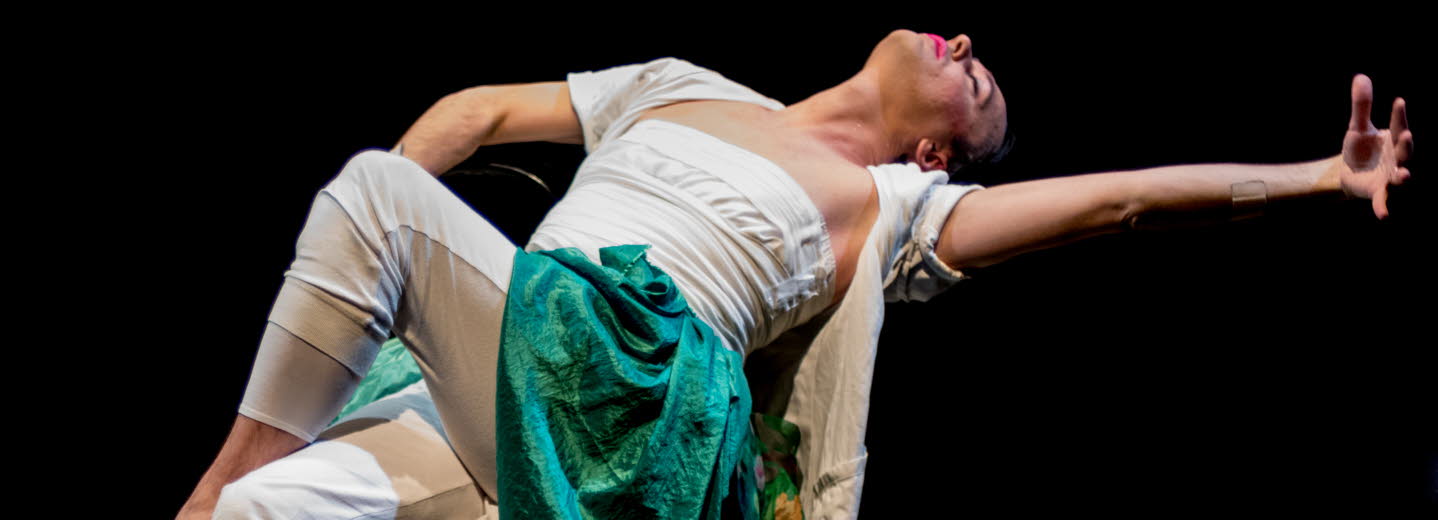 En dansare som sitter på en stol och gör en rörelse bakåt. Långa textiler som släpar på scengolvet. 