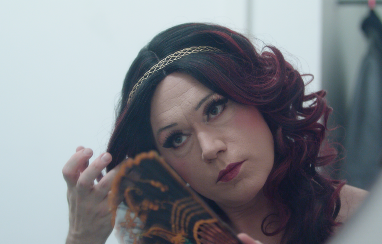 Närbild på uppsminkad dragqueen som håller i en solfjäder. Långt violettfärgat hår och guldigt diadem.