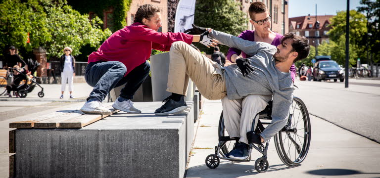 Tre dansare på ett soligt torg, där en av dansarna sitter i rullstol.