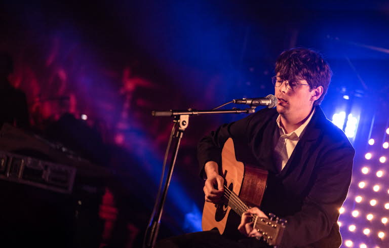 Ung man sitter på en scen, spelar gitarr och sjunger. 