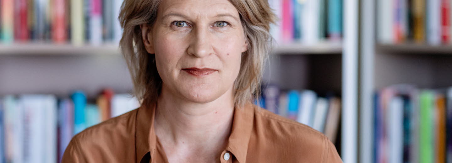 Medelålderskvinna i halvfigur. I bakgrunden bokhyllor fyllda med böcker. 