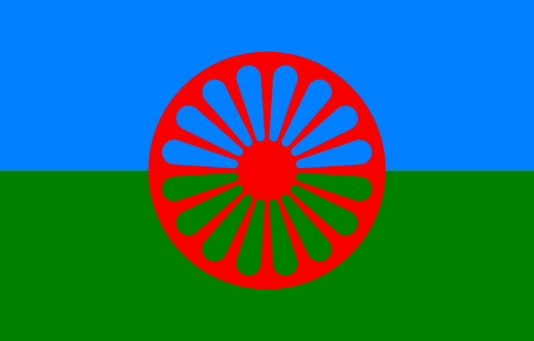 Romska flaggan, bakgrundens övre del är blå och den undre delen är grön. Framför finns röd ihålig cirkelformad symbol.