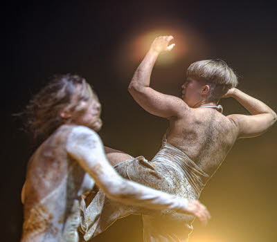 Två dansare med jordiga kläder, mörk bakgrund och en stark strålkastare. 