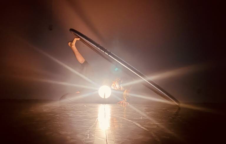 En kvinnlig dansare ligger på sidan i ett mörkt rum. Det ena benet är uppsträckt där foten balanserar en stor ring. Framför henne står en lampa som likt en strålkastare riktas mot betraktaren.