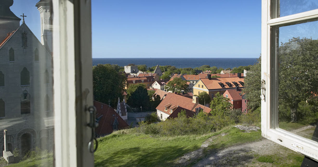 Ett öppet träfönster visar en vy över hustak, i horisonten syns havet som möter en blå himmel. Huset ligger högt och det syns lite berg och gräs precis utanför fönstret.