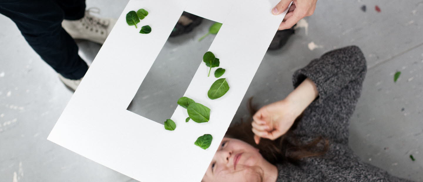 En kvinna ligger på rygg och tittar upp mot ett genombrutet papperark som hålls ovanför hennes huvud. Det ligger gröna blad på pappersarket. 