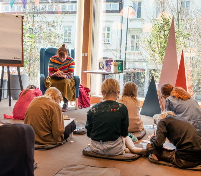 En grupp barn sitter utspridda framför en kvinna i en fåtölj, alla sitter böjda över sina skrivböcker.