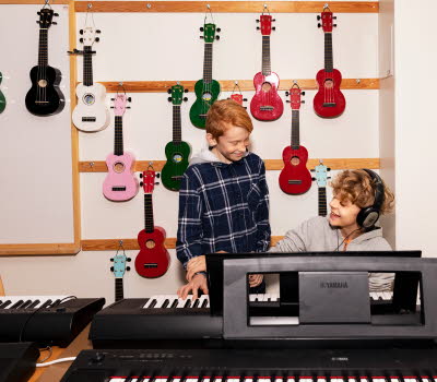 Två pojkar i musiksal, ena pojekn sitter vid ett piano, den andre står bredvid. Bakom pojkarna hänger flera mindre gitarrer i olika färger.