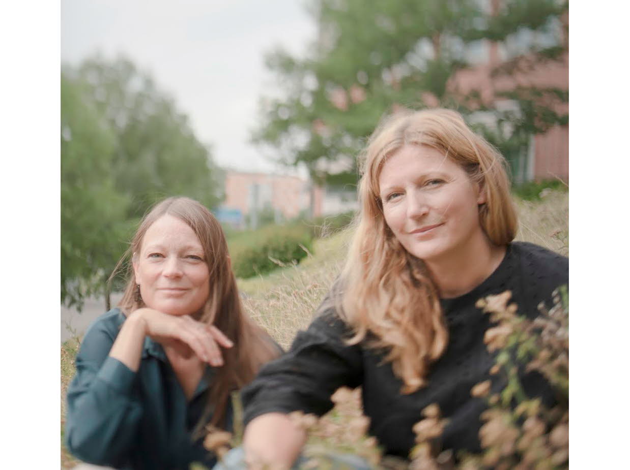 Foto på Jenny och Lotta som sitter ner i gräset, i bakgrunden skymtas träd och en tegelbyggnad.