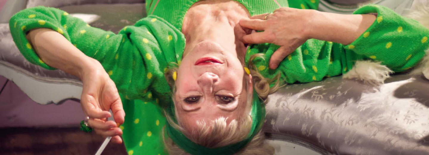 Book cover of "Vem tror hon att hon är": it's a photo of a women lies upside down on a couch wearing a green sweatsuit with green dots, smoking a cigarette.