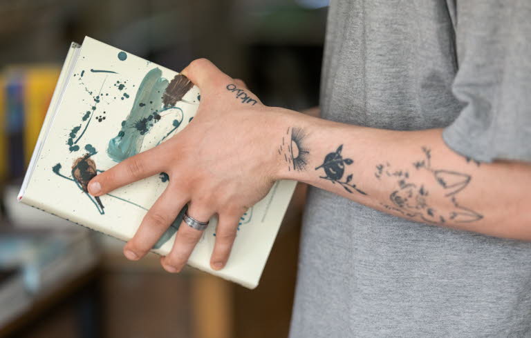 Närbild på tatuerad arm, handen håller i en bok.