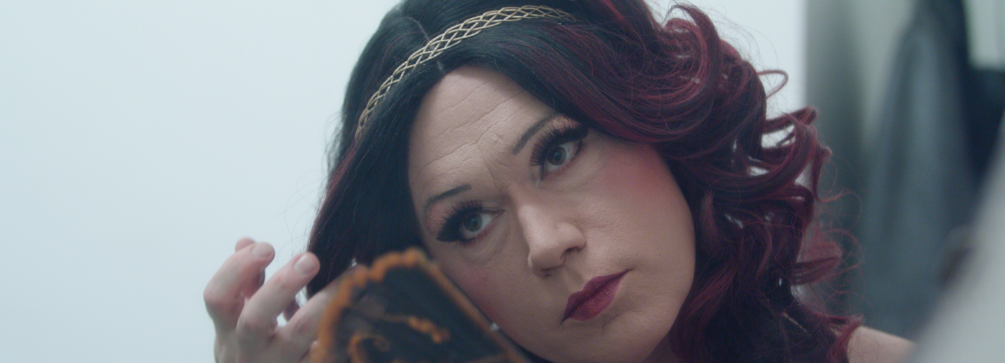 Närbild på uppsminkad dragqueen som håller i en solfjäder. Långt violettfärgat hår och guldigt diadem.