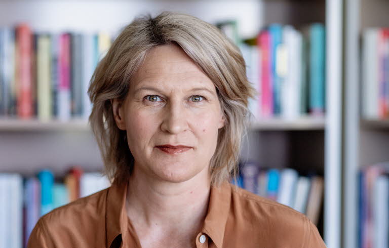 Medelålderskvinna i halvfigur. I bakgrunden bokhyllor fyllda med böcker. 