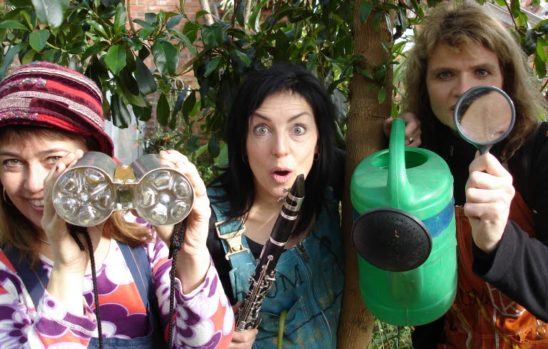 Tre personer står bland träd och tittar nyfiket in i kameran. De håller i en vattenkanna, en flöjt och en kikare.