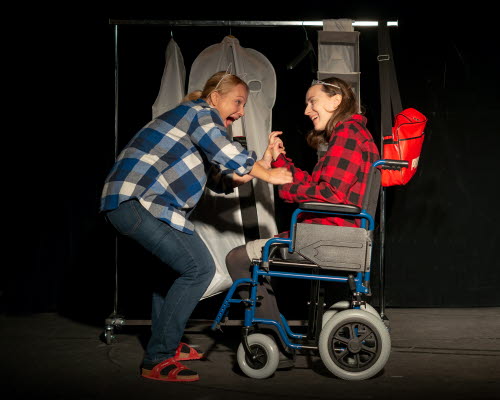 Två kvinnor på en scen spexar och skrattar tillsammans. Den ena är rullstolsburen.