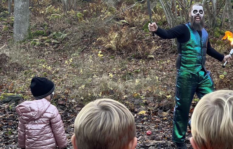 Barn som tittar på en artist i grönblank kostym med en fackla i handen ute i skogen. 