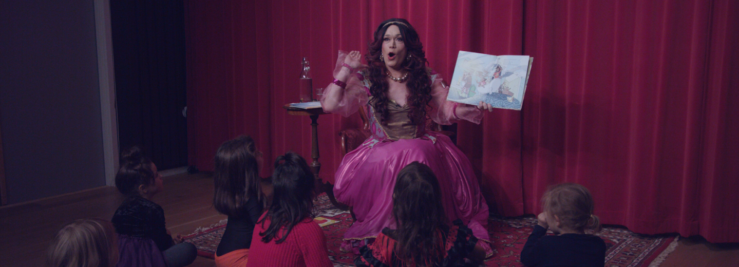 En dragqueen som läser högt för en grupp barn som sitter på golvet. 