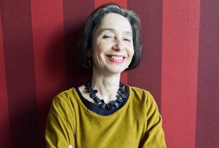 Foto på Elena Balzamo som ler mot kameran. Elena har en mörk page och är klädd i en senapsgul klänning med ett stort svart halsband. Bakgrunden är randig i röda toner. 