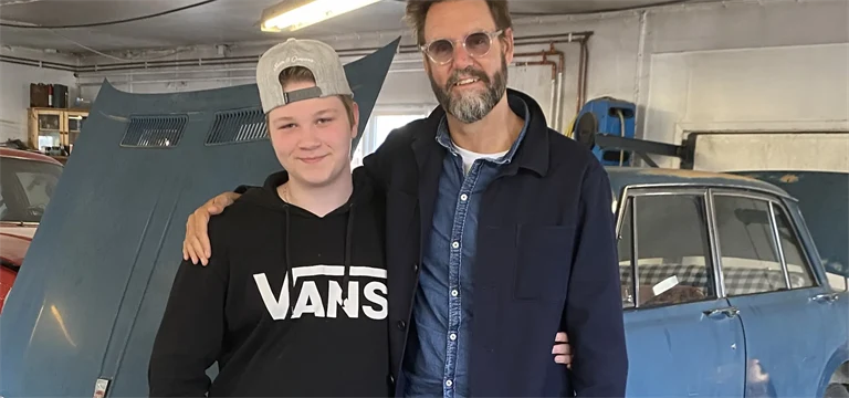 En ung kille står tillsammans med en författare framför en bil med öppen motorhuv.