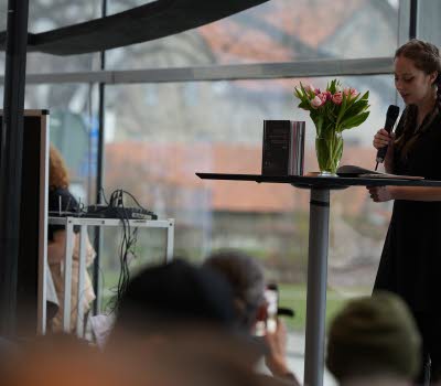 En ung kvinna står med en mikrofon på en scen och läser ur en bok.