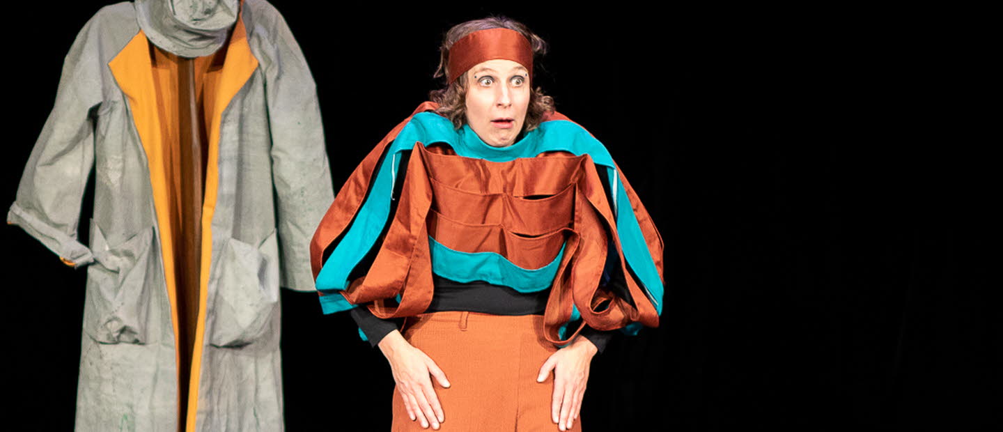 Två kvinnor på scen. En spelar på keyboard som ligger på ett figurliknande träbord och den andra tittar storögt mot publiken, axlarna är upphöjda. Båda har färgstarka kläder i turkost och ockra. I bakgrunden hänger en grå rock med tillhörande hatt.