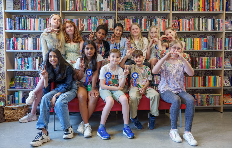 Grupp ungdomar i blandade åldrar sitter i två rader framför en stor bokhylla. De ser glada ut och har årets Väck boken-rosett på sig.