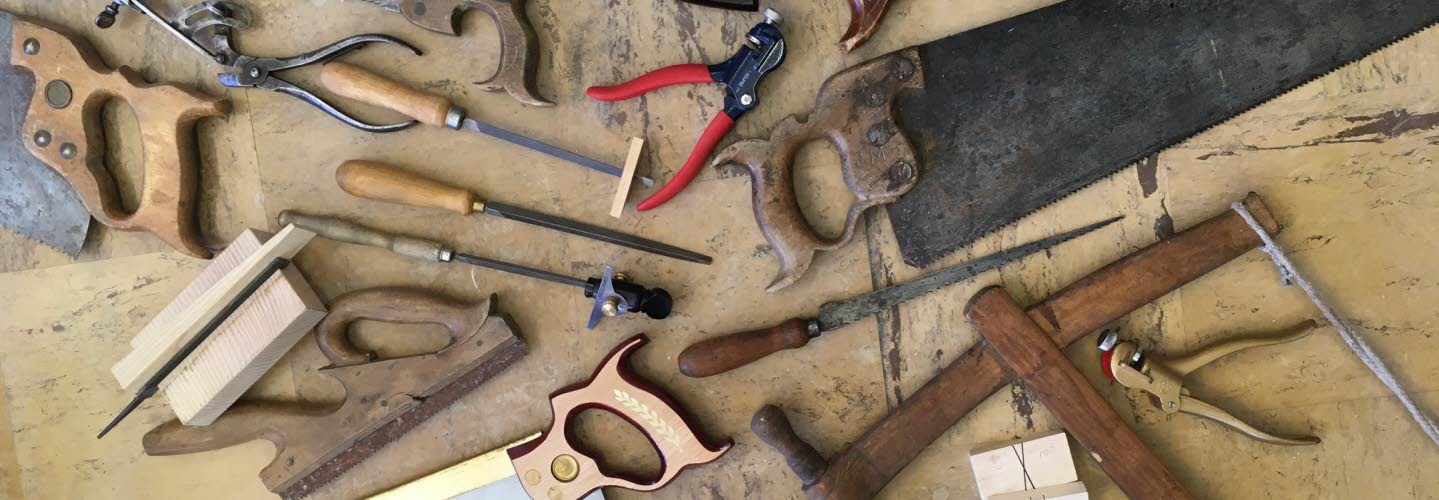 Gamla och nya handverktyg, främst sågar, filar och tänger, utspridda på ett slitet träbord.