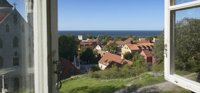 Ett öppet träfönster visar en vy över hustak, i horisonten syns havet som möter en blå himmel. Huset ligger högt och det syns lite berg och gräs precis utanför fönstret.