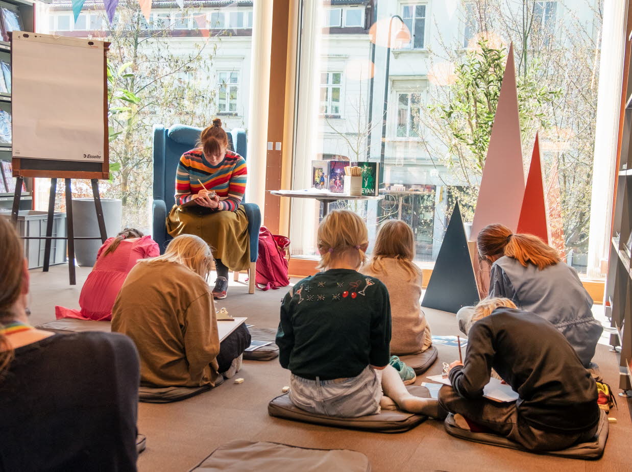 En grupp barn sitter utspridda framför en kvinna i en fåtölj, alla sitter böjda över sina skrivböcker.