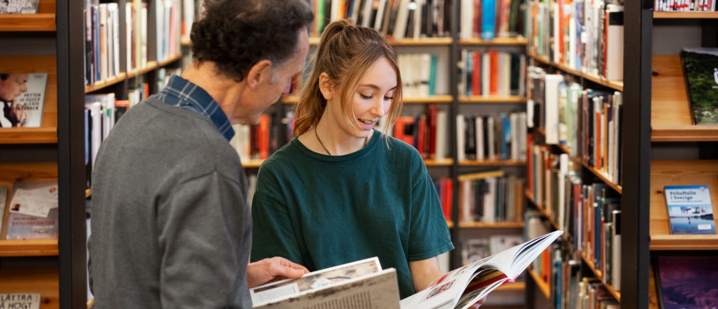 Två bibliotekarier står bland bokhyllor, bläddrar i varsin bok och diskuterar innehållet. 