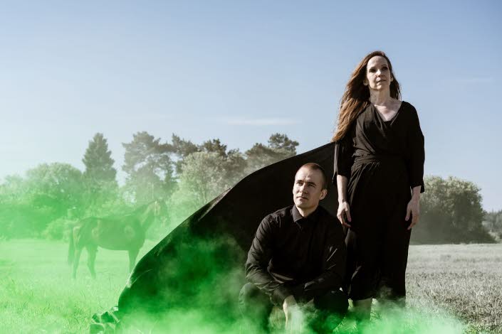 En man och en kvinna i svart klädsel på ett torrt gräsfält med grön rök.