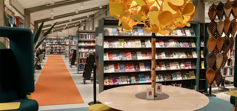 Interiör från bibliotek med bokhyllor, sittmöbler och samlingsbord med en lampa som ser ut som ett träd.