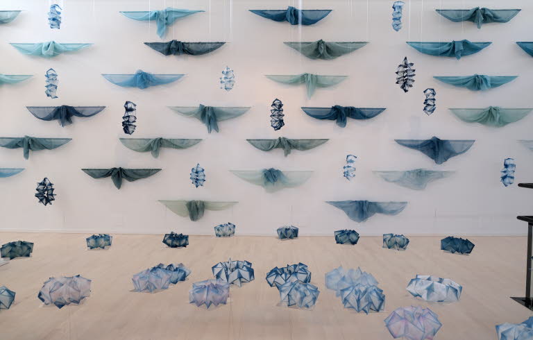 Konstinstallation med blå och gröna tygstycken upphängda i rader på en vit vägg, samt geometriska former i liknande färger placerade på golvet.