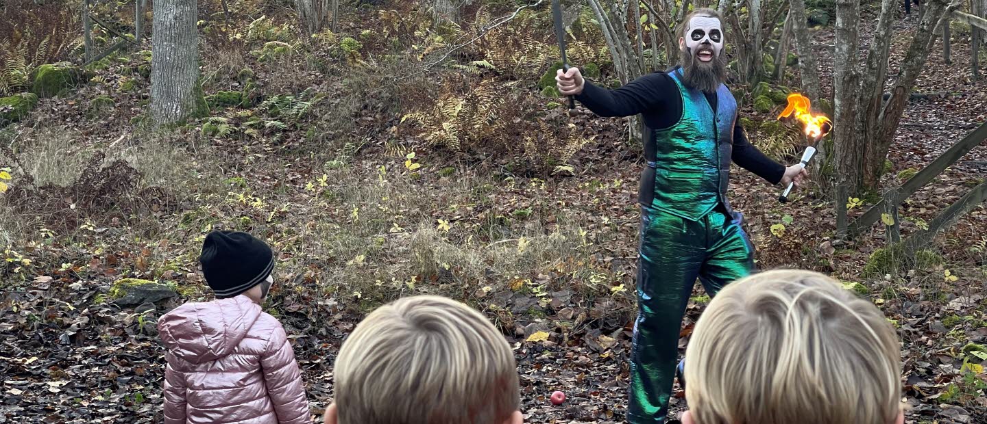 Barn som tittar på en artist i grönblank kostym med en fackla i handen ute i skogen. 